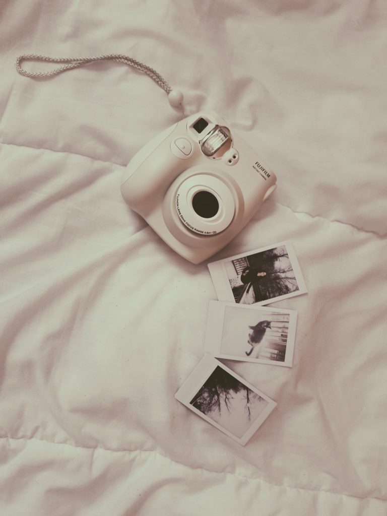 O camera foto instant asezata pe pat langa cateva fotografii de asemenea instant 