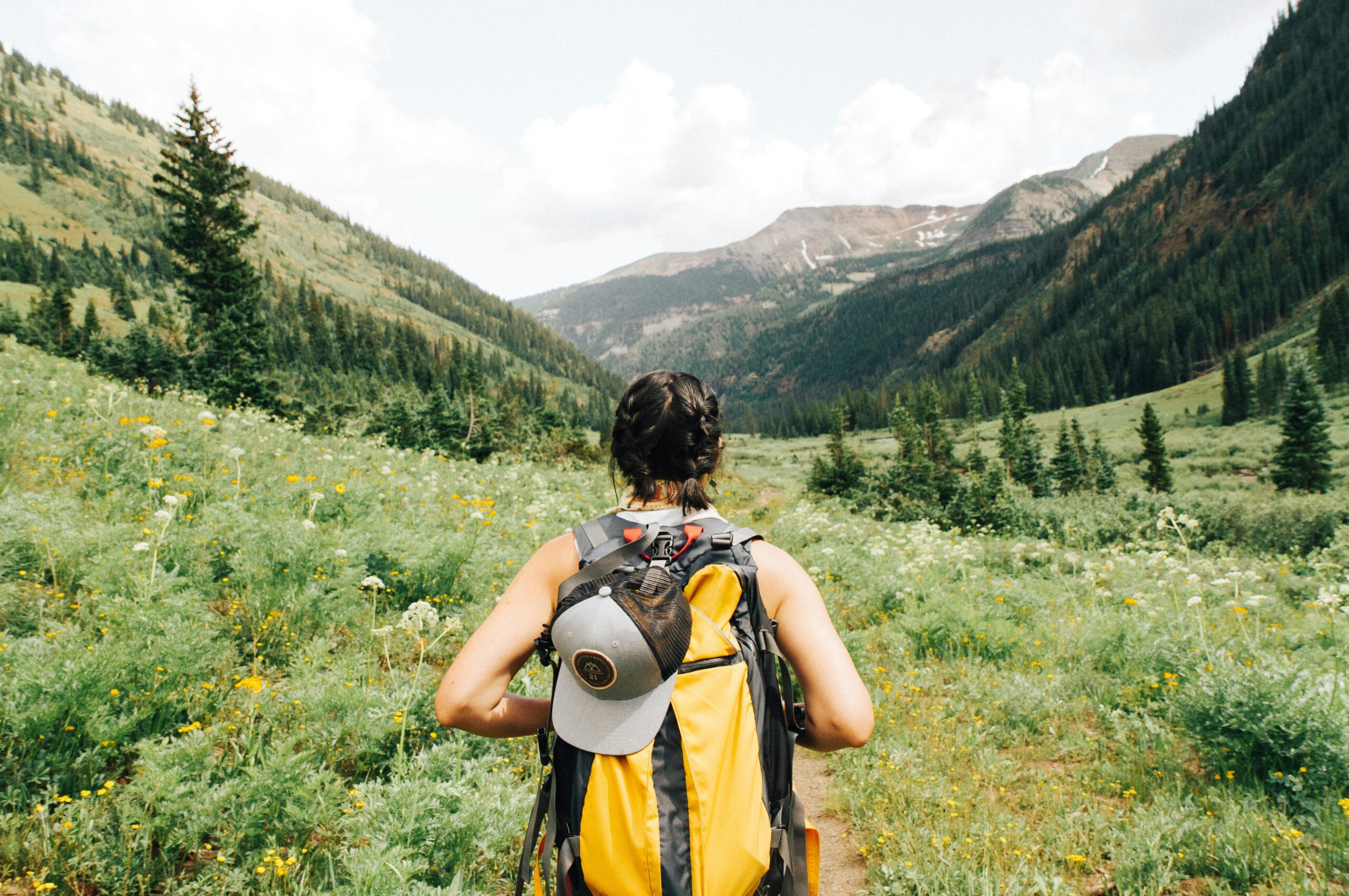 O fată în drumeție pe munte, echipată sport, cu toate lucrurile esențiale pentru un traseu, este înconjurată de natură, brazi și munți.