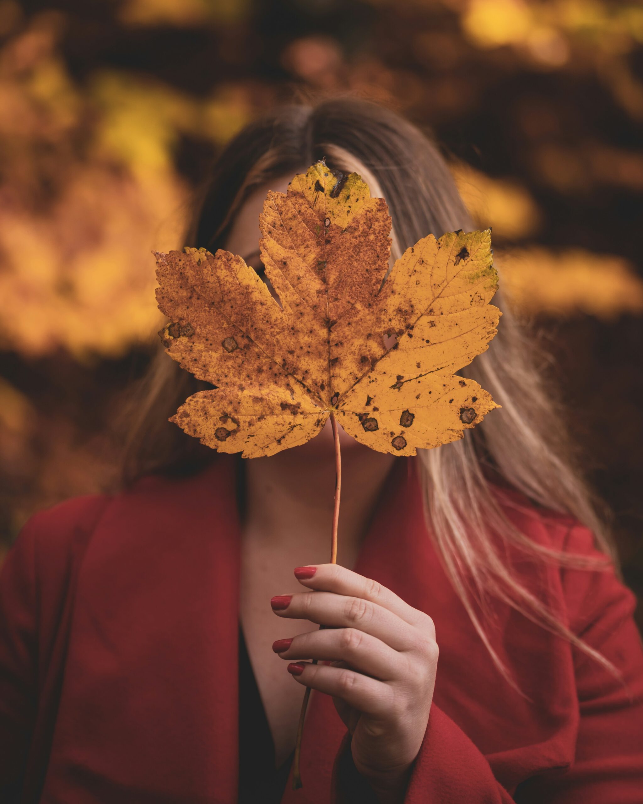 o poza cu o fata imbracata cu o haina rosie ce se asorteaza cu unghiile acesteia. Fata pozeaza pentru o fotografie foarte populara de toamna, in care isi ascunde fata in spatele unei frunze galbene